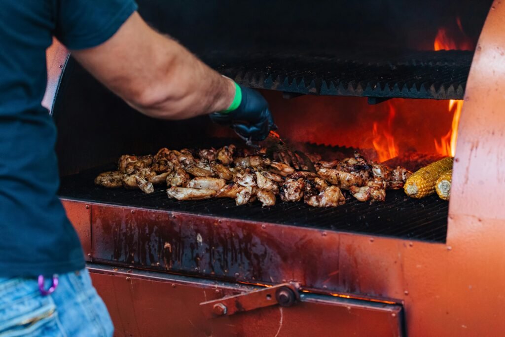 Close-up of grilling chicken wings and corn on an outdoor barbecue with flames.